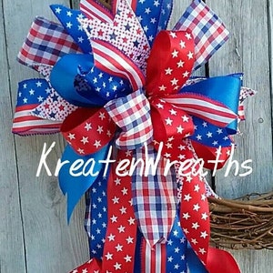 May include: A red, white, and blue patriotic wreath bow with a variety of patterned ribbons, including stars, stripes, and gingham. The bow is tied with a blue ribbon and is attached to a grapevine wreath.