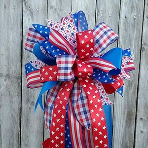 May include: A red, white, and blue bow made with patterned ribbon, perfect for a patriotic display. The bow is tied around a black lantern with a decorative design.