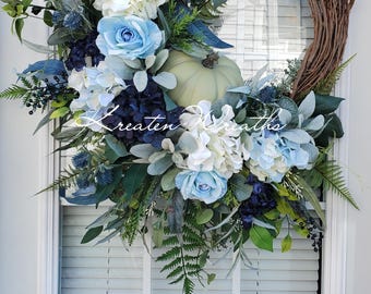 Può includere: Una ghirlanda di rami di vite con rose azzurre e bianche, ortensie blu scuro, fogliame verde salvia, felci e una zucca verde pallido al centro. Il testo Kreaten Wreaths è sovrapposto in bianco in corsivo al centro della composizione.