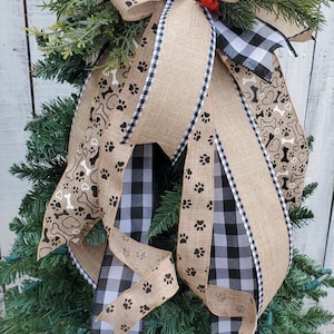 Pode incluir: Um laço para o topo da árvore de Natal com uma fita de juta e xadrez preto e branco. O laço é decorado com desenhos de pegadas e ossos, bagas vermelhas e um sino marrom.