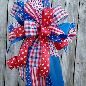 May include: A red, white, and blue patriotic bow made with various patterned ribbons, including gingham, stars, and stripes. The bow is tied around a black lantern with a gold patterned design. The bow is sitting on a white surface with two white daisies.