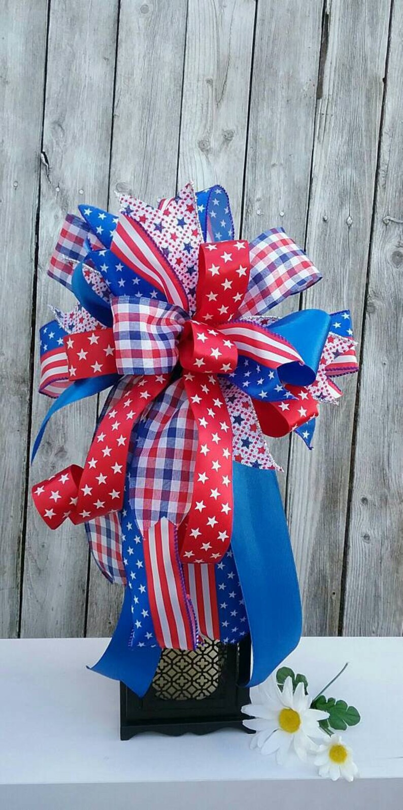 May include: A red, white, and blue patriotic bow made with various patterned ribbons, including gingham, stars, and stripes. The bow is tied around a black lantern with a gold patterned design. The bow is sitting on a white surface with two white daisies.