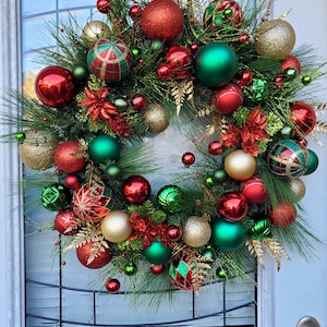 May include: A Christmas wreath decorated with red, green, and gold ornaments. The wreath is made of pine branches and has a variety of different ornaments, including balls, stars, and bells.