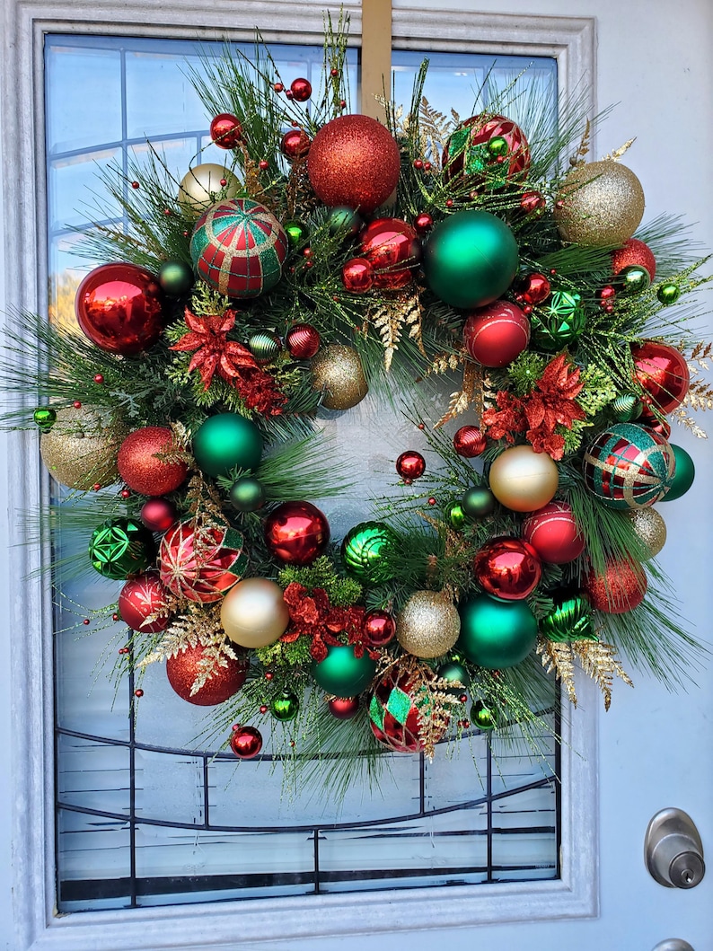 May include: A Christmas wreath decorated with red, green, and gold ornaments. The wreath is made of pine branches and has a variety of different ornaments, including balls, stars, and bells.
