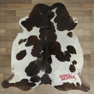 May include: A cowhide rug with a natural pattern of brown and white spots. The rug has an irregular shape and is designed to be placed on a floor. The rug has the text "Hides Bazaar" in red.