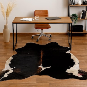 May include: A black and white cowhide rug with a brown center is on a hardwood floor. A wooden desk with a black metal frame, a brown leather chair, and a bookshelf are in the background. A vase with dried plants is on the left.