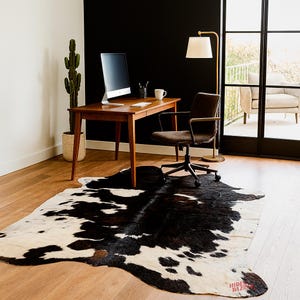 May include: A black and white cowhide rug in an office setting. The rug is placed under a wooden desk with a computer monitor, a brown office chair, and a gold floor lamp. A cactus in a woven pot is in the corner.