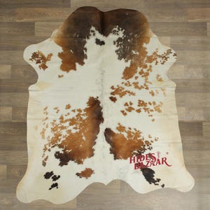 May include: A cowhide rug with a natural pattern of brown and white spots. The rug has an irregular shape and is laid out on a wooden floor. The logo "Hides Bazaar" is visible in the bottom right corner.