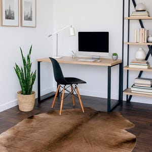 May include: A modern home office setup featuring a light wood desk with black metal legs, a black chair with wooden legs, and a cowhide rug. A computer monitor, desk lamp, and framed art pieces complete the space.
