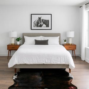 May include: A bedroom scene featuring a bed with a gray headboard, white bedding, and a dark gray lumbar pillow. Two wooden nightstands with lamps flank the bed. A black and white horse print hangs above the bed, and a cowhide rug is on the floor.