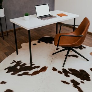 May include: A modern home office setup featuring a white desk with black legs, a brown leather chair, and a laptop. A cowhide rug with brown and white patches covers the floor. A small potted plant and a notebook are on the desk.