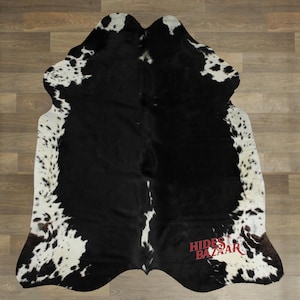 May include: A cowhide rug with a black and white spotted pattern. The rug has a natural shape and texture, with the brand name "Hides Bazaar" in red text. The rug is displayed on a wooden floor.