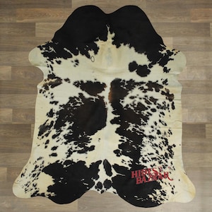 May include: A cowhide rug with a natural black and white spotted pattern. The rug has a unique shape and is laid out on a wooden floor. The rug has the text "HIDES BAZAAR" in red.