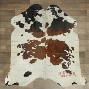 May include: A cowhide rug with a natural pattern of black, brown, and white spots. The rug has an irregular shape and is laid on a wooden floor. The text "HIDE BAZAAR" is visible in the lower right corner.
