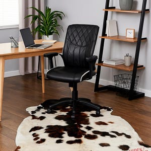 May include: A black and white cowhide rug with brown spots is on a hardwood floor. A black leather office chair with white trim sits above the rug. A wooden desk with a laptop and a black metal and wood shelf unit are in the background.