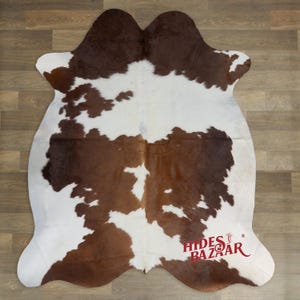 May include: A cowhide rug with a brown and white pattern. The rug has an irregular shape and is made of natural leather. The logo "Hides Bazaar" is visible in the bottom right corner.