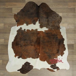 May include: A cowhide rug with a natural pattern of brown and white patches. The rug has an irregular shape and is laid on a wooden floor. The rug is a decorative item for home decor.