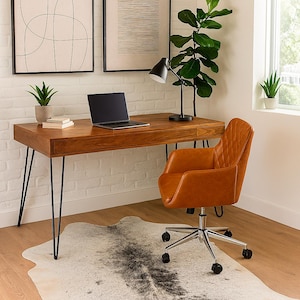 May include: A home office setup featuring a wooden desk with black hairpin legs, a laptop, and a black desk lamp. An orange leather office chair sits on a cowhide rug. Two framed abstract art pieces hang on the white brick wall.