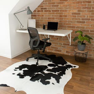 May include: A modern home office setup featuring a black and white cowhide rug, a black mesh office chair, and a white desk with a laptop. The room has a brick wall and a gray desk lamp.