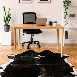 May include: A modern home office setup featuring a light wood desk with a laptop, black office chair, and a black and white cowhide rug. Two framed landscape prints hang above the desk, with a tall plant and a white shelf unit in the background.
