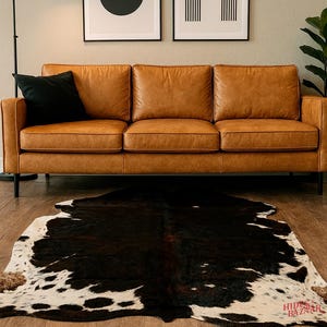 May include: A brown leather sofa with a black pillow sits above a cowhide rug with black and white patterns. The rug is on a wooden floor. Two framed abstract art pieces hang on the wall behind the sofa. A black lamp is to the left.