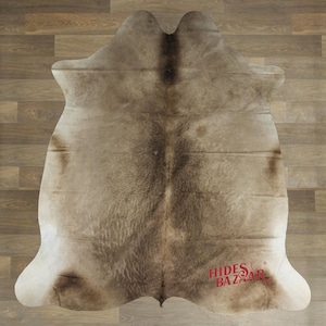 May include: A cowhide rug with a natural color palette of beige, brown, and cream. The rug has a unique shape and texture, with a central dark brown stripe and lighter edges. The rug is a decorative floor covering.