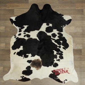 May include: A cowhide rug with a black and white spotted pattern. The rug has a natural shape and a brown patch near the center. The logo "Hides Bazaar" is visible in the bottom right corner. The rug is placed on a wooden floor.