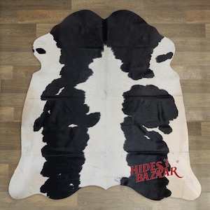 May include: A cowhide rug with a distinctive black and white pattern. The rug has irregular edges and a natural shape, with large patches of black against a white background. The logo "Hides Bazaar" is visible in the lower right corner.