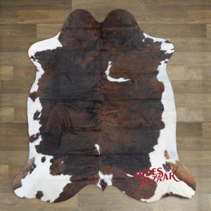 May include: A cowhide rug with a natural pattern of brown and white patches. The rug has an irregular shape, with a dark brown center and white edges. The rug is placed on a wooden floor.