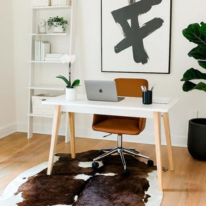 May include: A modern home office setup featuring a white desk with wooden legs, a brown leather office chair, and a laptop. A cowhide rug in brown and white sits beneath the desk. A large abstract art piece hangs on the wall.
