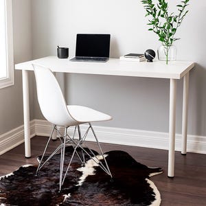 May include: A modern home office setup featuring a white desk with a laptop, black mug, and decorative items. A white chair with chrome legs sits on a cowhide rug. A glass vase with greenery adds a natural touch to the workspace.