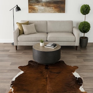 May include: A living room scene featuring a brown and white cowhide rug on a wood floor. A gray sofa with throw pillows sits against a white wall, with a round coffee table and abstract art. A black floor lamp and potted topiary complete the decor.