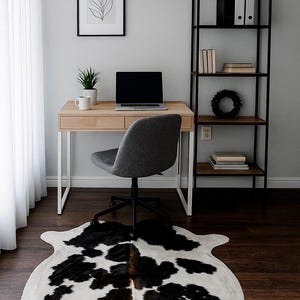 May include: A modern home office setup featuring a light wood desk with a laptop, a gray chair, and a black metal bookshelf. A black and white cowhide rug covers the dark wood floor. A framed botanical print hangs on the wall.