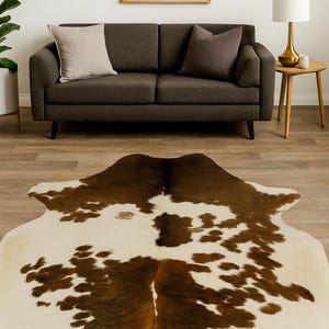 May include: A cowhide rug with a brown and white spotted pattern. The rug is irregularly shaped and lies on a hardwood floor. A dark gray sofa with two pillows is in the background.