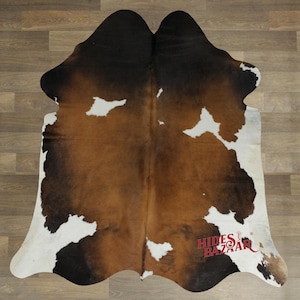 May include: A cowhide rug with a natural pattern. The rug features a mix of brown, black, and white patches. The rug is displayed on a wooden floor. The text "Hides Bazaar" is visible in the lower right corner.