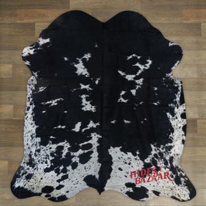 May include: A cowhide rug with a black and white spotted pattern. The rug has an irregular shape and is made of natural materials. The rug is on a wooden floor. The text "HIDES BAZAAR" is visible in red.