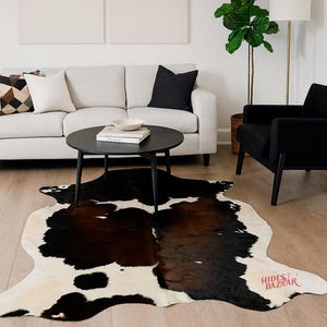 May include: A cowhide rug with a distinctive pattern of black and brown patches on a cream background. The rug is placed on a hardwood floor, with a black round coffee table and a light-colored sofa in the background. The rug's shape is irregular.