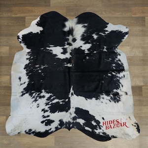 May include: A cowhide rug with a black and white spotted pattern. The rug has an irregular shape and is made of natural animal hide. The rug is placed on a wooden floor.