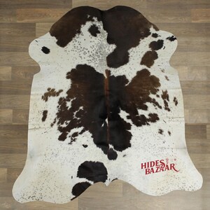 May include: Cowhide rug with a natural pattern of white, brown, and black spots. The rug has an irregular shape and a soft texture. The brand name "Hides Bazaar" is printed in red near the bottom right corner. The rug is placed on a wooden floor.
