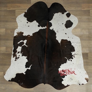 May include: Cowhide rug with a natural pattern of black and white spots. The rug has an irregular shape and a brown stripe down the center. The rug is made of genuine cowhide and is suitable for home decor.