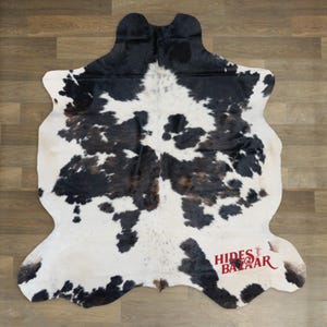 May include: Cowhide rug with a natural pattern of black and brown spots on a white background. The rug has an irregular shape and is likely made of genuine cowhide. The rug is displayed on a wooden floor.