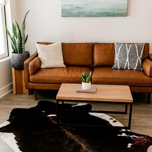 May include: A living room scene featuring a brown leather sofa with two decorative pillows, a wooden coffee table, and a cowhide rug. A painting of a seascape hangs above the sofa. A potted plant sits on a wooden stand.