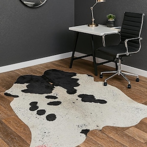 May include: A cowhide rug with a black and white spotted pattern. The rug is on a hardwood floor in a room with a desk, chair, and mirror. The rug has a unique shape and adds a rustic touch to the space.