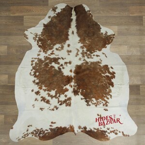 May include: A cowhide rug with a natural pattern of brown and white spots. The rug has an irregular shape and is likely made of genuine leather. The logo "Hides Bazaar" is visible in the bottom right corner.