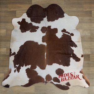 May include: A cowhide rug with a brown and white pattern. The rug has an irregular shape and is made of natural materials. The rug is displayed on a wooden floor. The logo "Hides Bazaar" is visible in the bottom right corner.