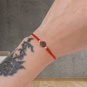 May include: A red string bracelet with a dark gray circular stone and silver beads. The bracelet is tied with a sliding knot closure. The bracelet is worn on a wrist.