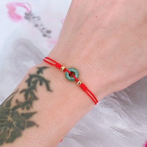 May include: A red string bracelet featuring a jade donut pendant and gold-colored beads. The jade is a light green color. The bracelet is tied with a sliding knot for adjustable sizing.