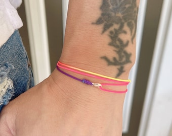 May include: A colorful friendship bracelet with three strands of string in shades of pink, yellow, and purple. The bracelet is tied with a silver charm.