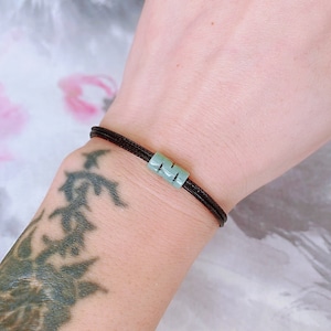 May include: A black string bracelet with a light green jade bead charm. The bead is square shaped and has a subtle pattern.