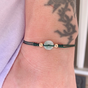 May include: A green string bracelet featuring a round, pale green stone and two small gold beads. The bracelet is tied with a dark green cord. The bracelet is a delicate piece of jewelry.